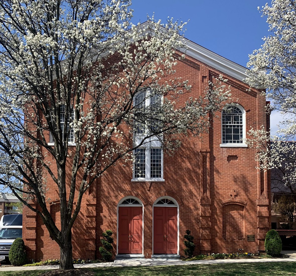 The historic 1855 Alfred Street Baptist Church building in Alexandria, Virginia: a red brick facade with twin red doors and a flowering tree in front.